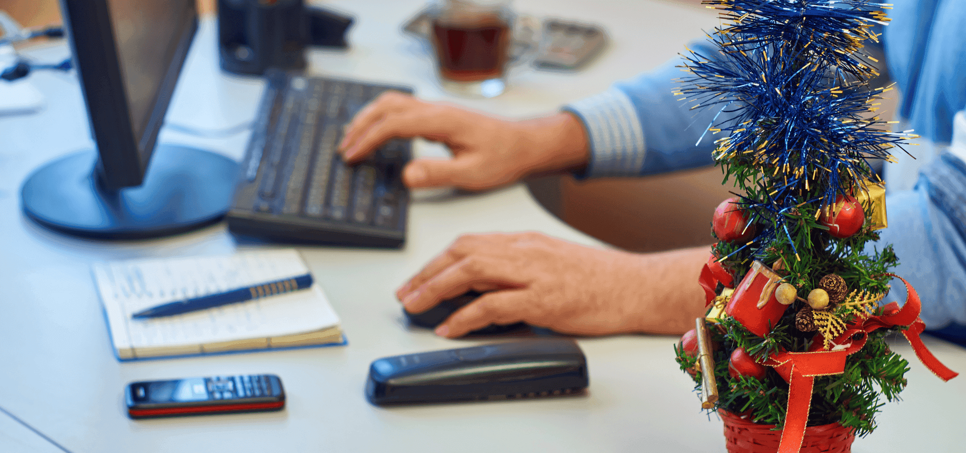Person arbeitet am Schreibtisch mit Computer, Notizen und Telefon, daneben ein kleiner weihnachtlich geschmückter Tannenbaum – Symbolbild für Arbeitsalltag in der Adventszeit