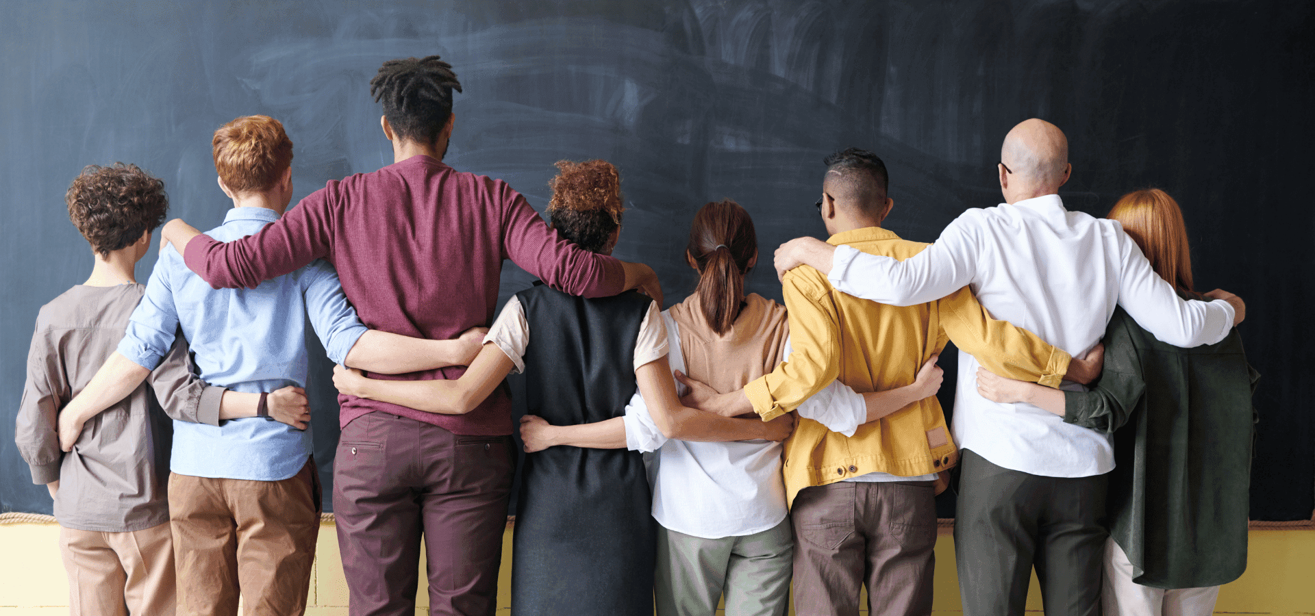 Group of people of different background standing with their back, arm in arm in front a wall.