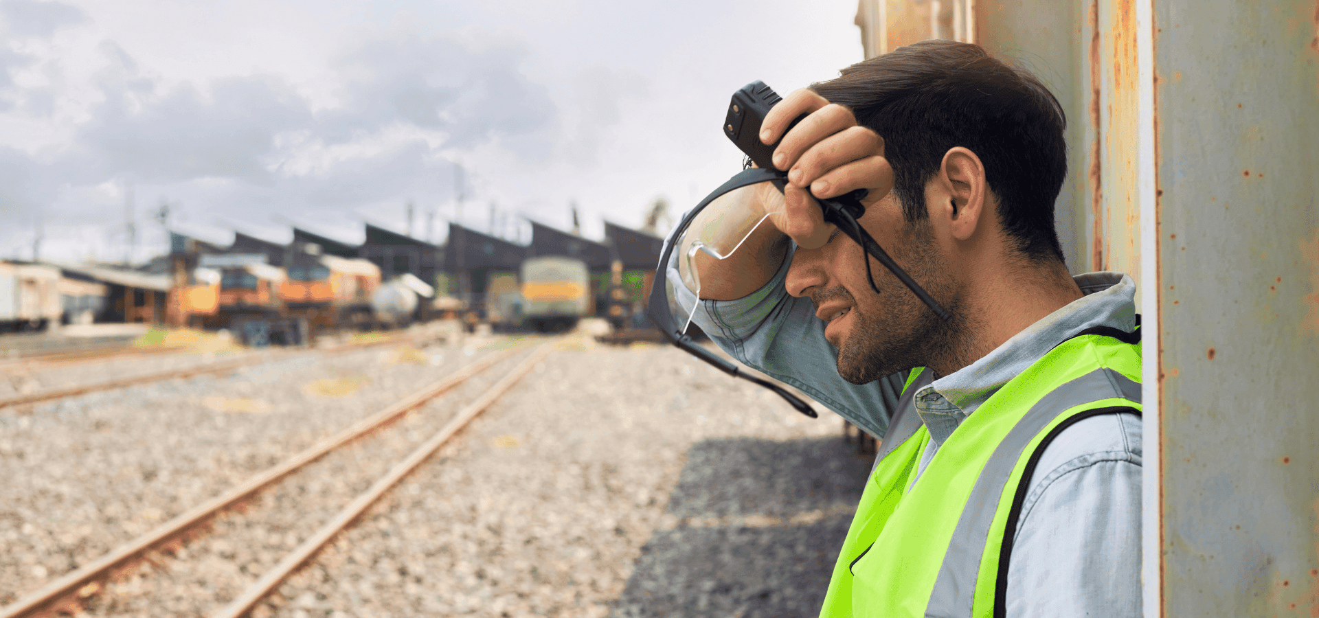 Mann mit Sicherheitsweste auf Bahnhof auf Gleisen, wischt sich mit Hand Schweiß von der Stirn
