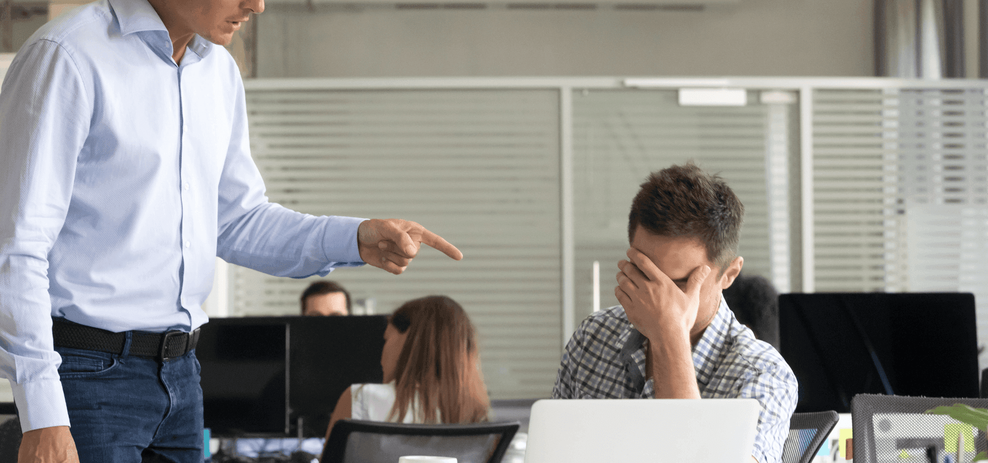 Boss screaming to employee, pointing to him while the employee is seated and his face sinks to his hands.