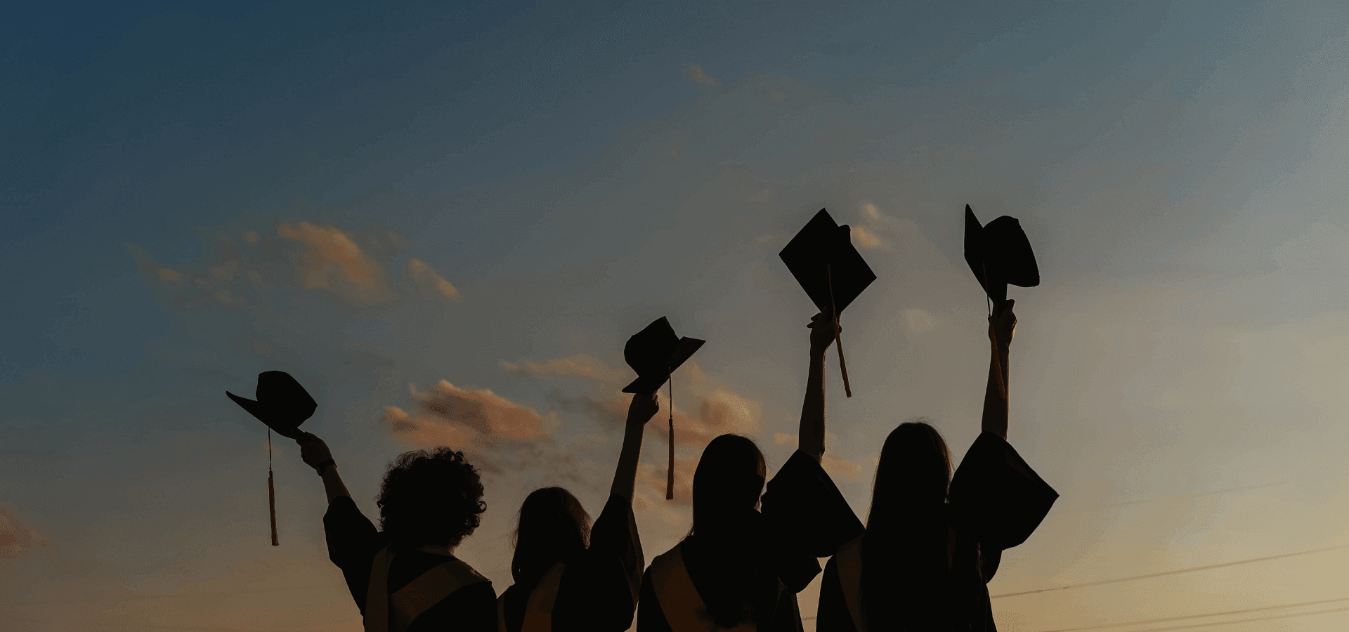 Graduating students holding up their hats in front a yellow-blue evening sky.