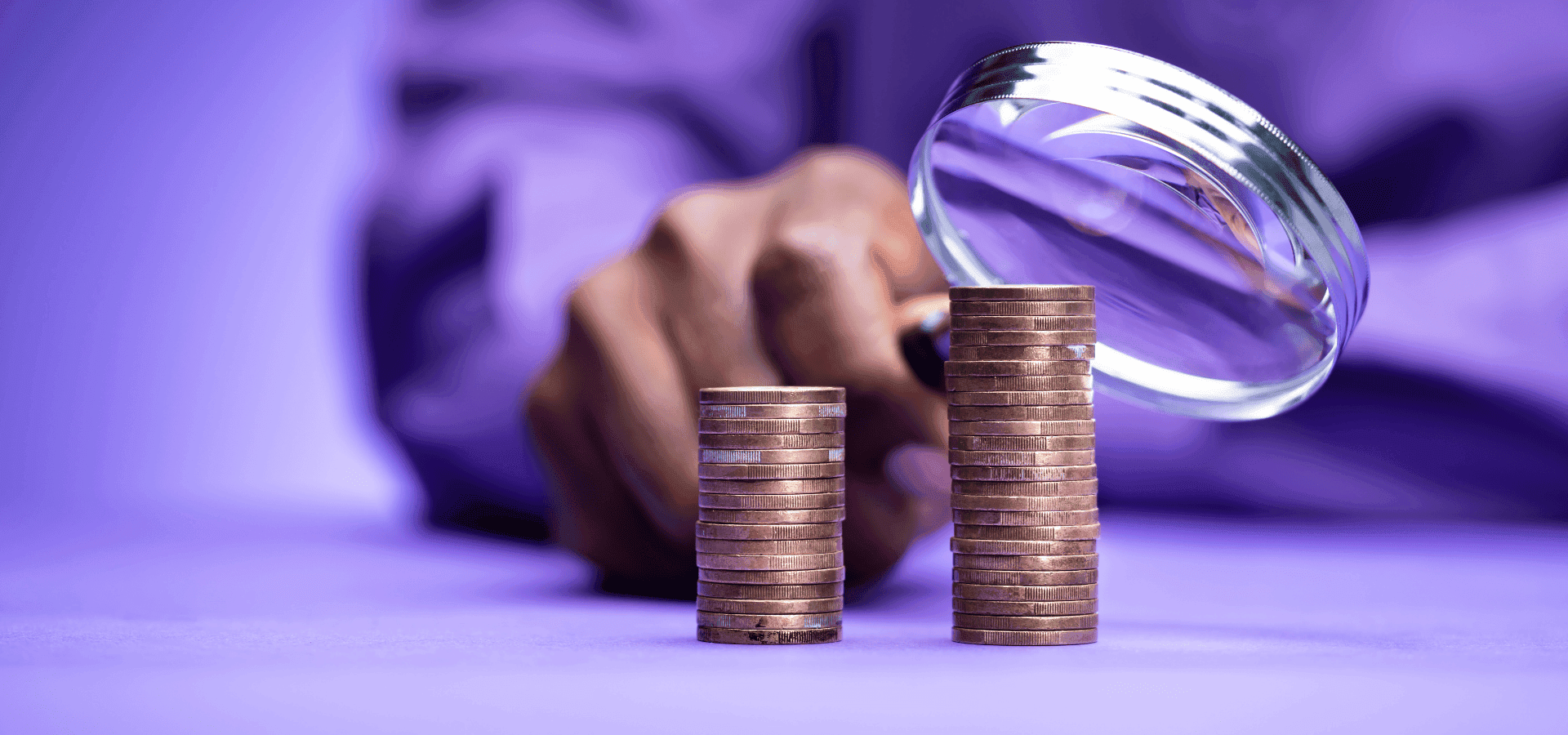 A hand holds a magnifying glass over two stacks of Euro coins of different heights, symbolizing wage or wealth analysis.
