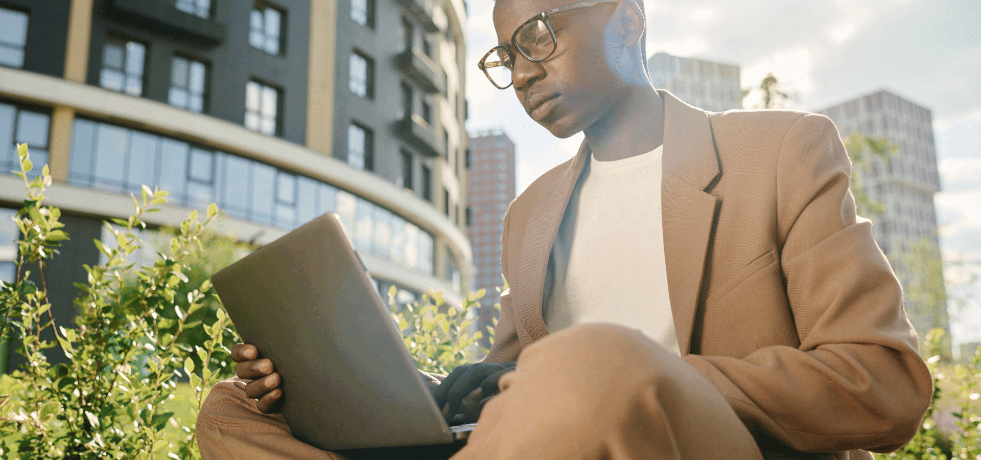 Young office worker sitting with his laptop in his lap in a green park in a CBD.